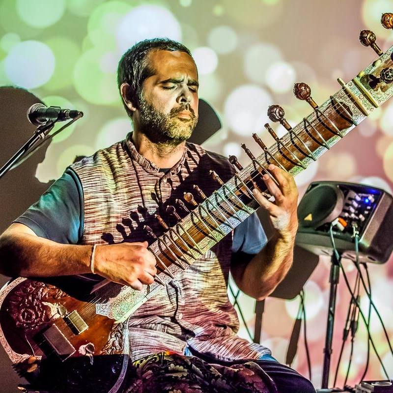 22539716_1607273576000614_2035492282134918566_n-1 Man playing sitar on colorful stage.