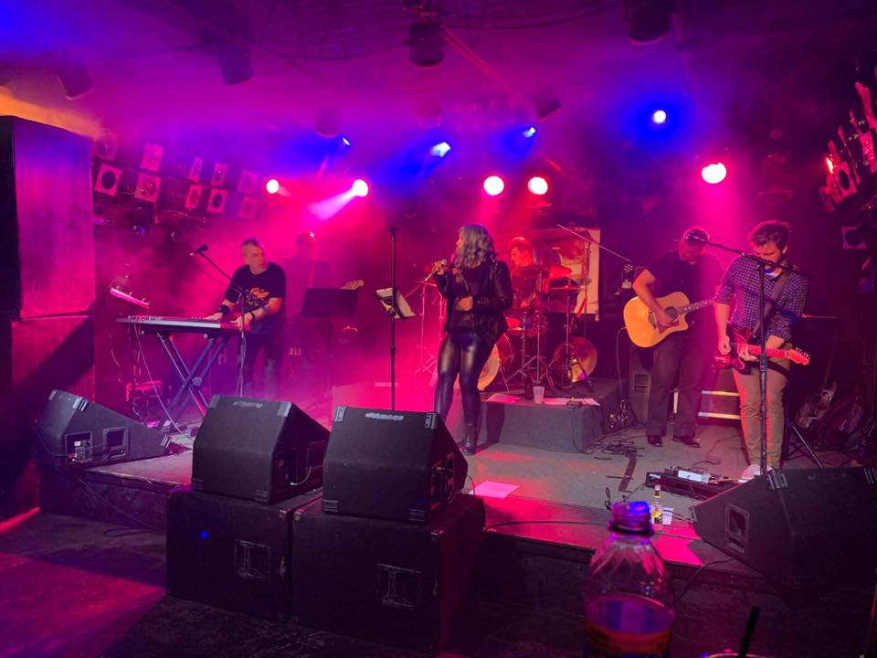 RG_CINEMATICS_MAY31st-1 Band performing on stage with colorful lights.